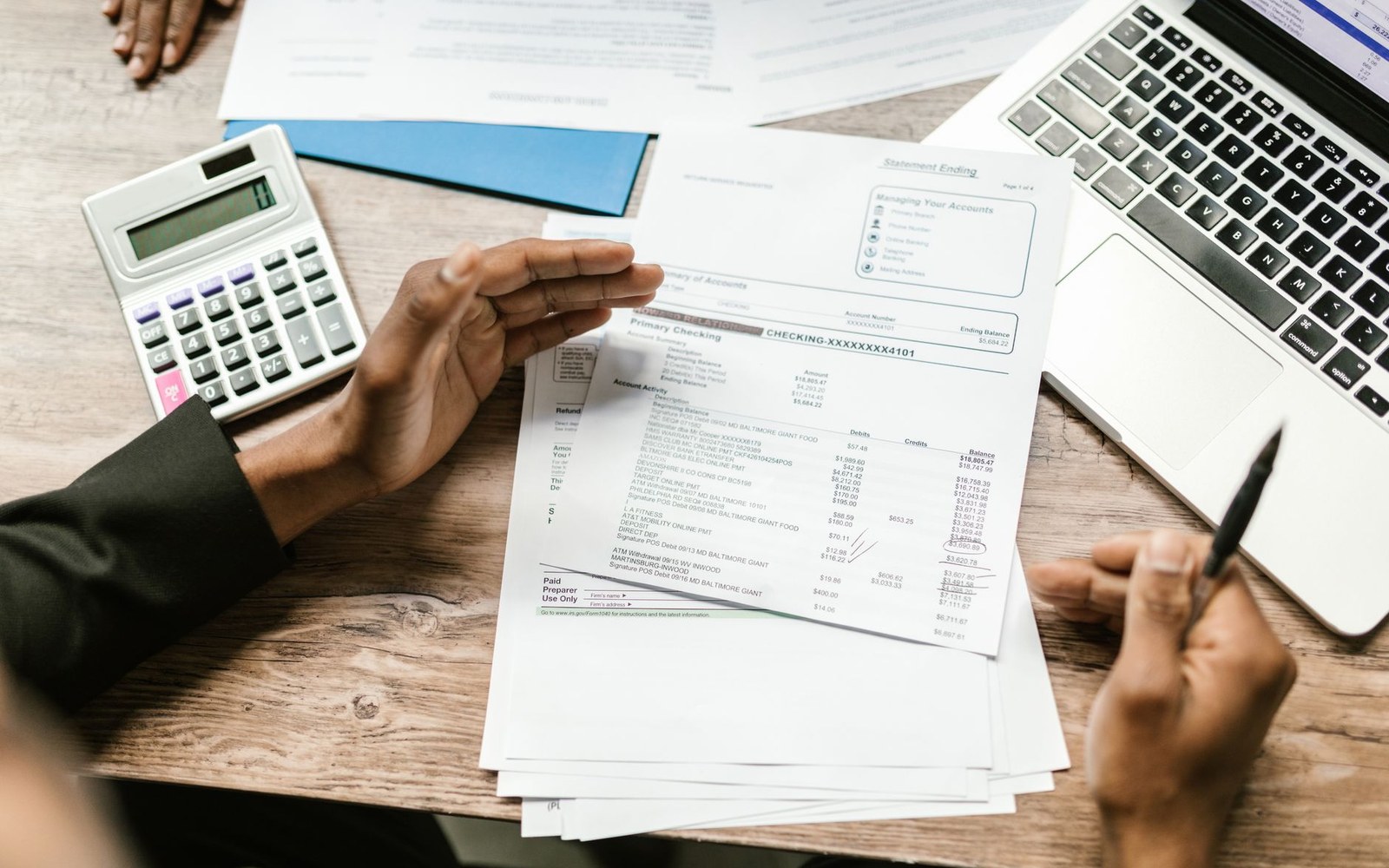 A bank statement face-down beside a pocket calculator on a plain desk, one hand resting on each, natural light