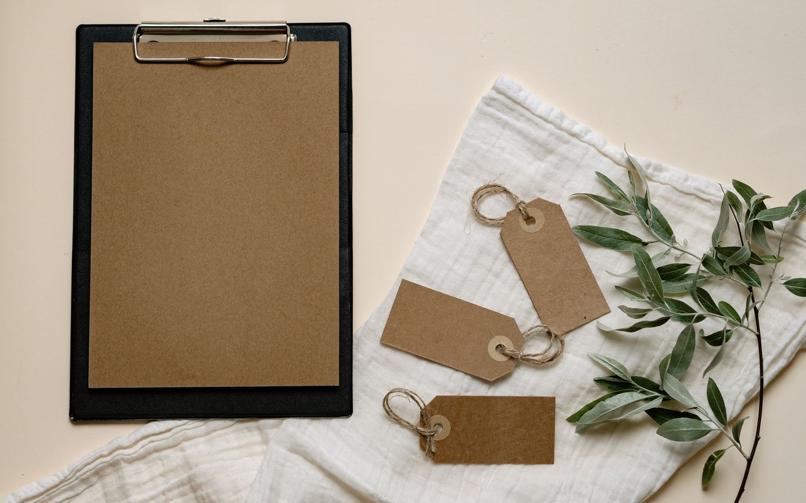 Blank clipboard and kraft paper gift tags arranged on a linen cloth with olive leaves