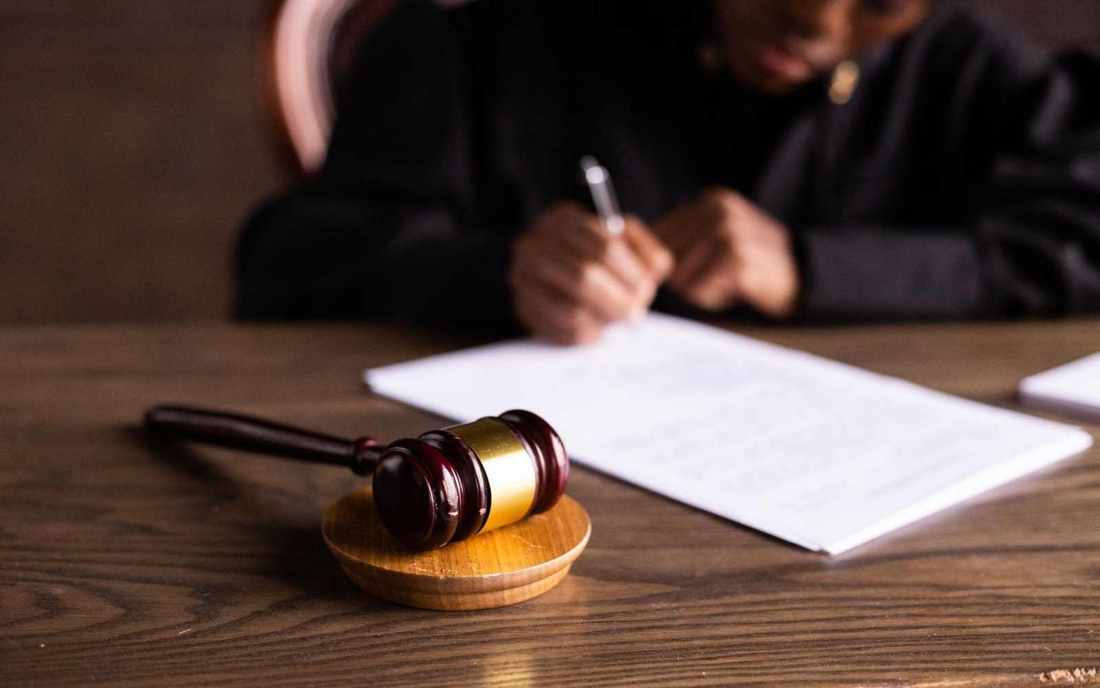 A wooden gavel rests on a desk while a robed figure signs a document behind it