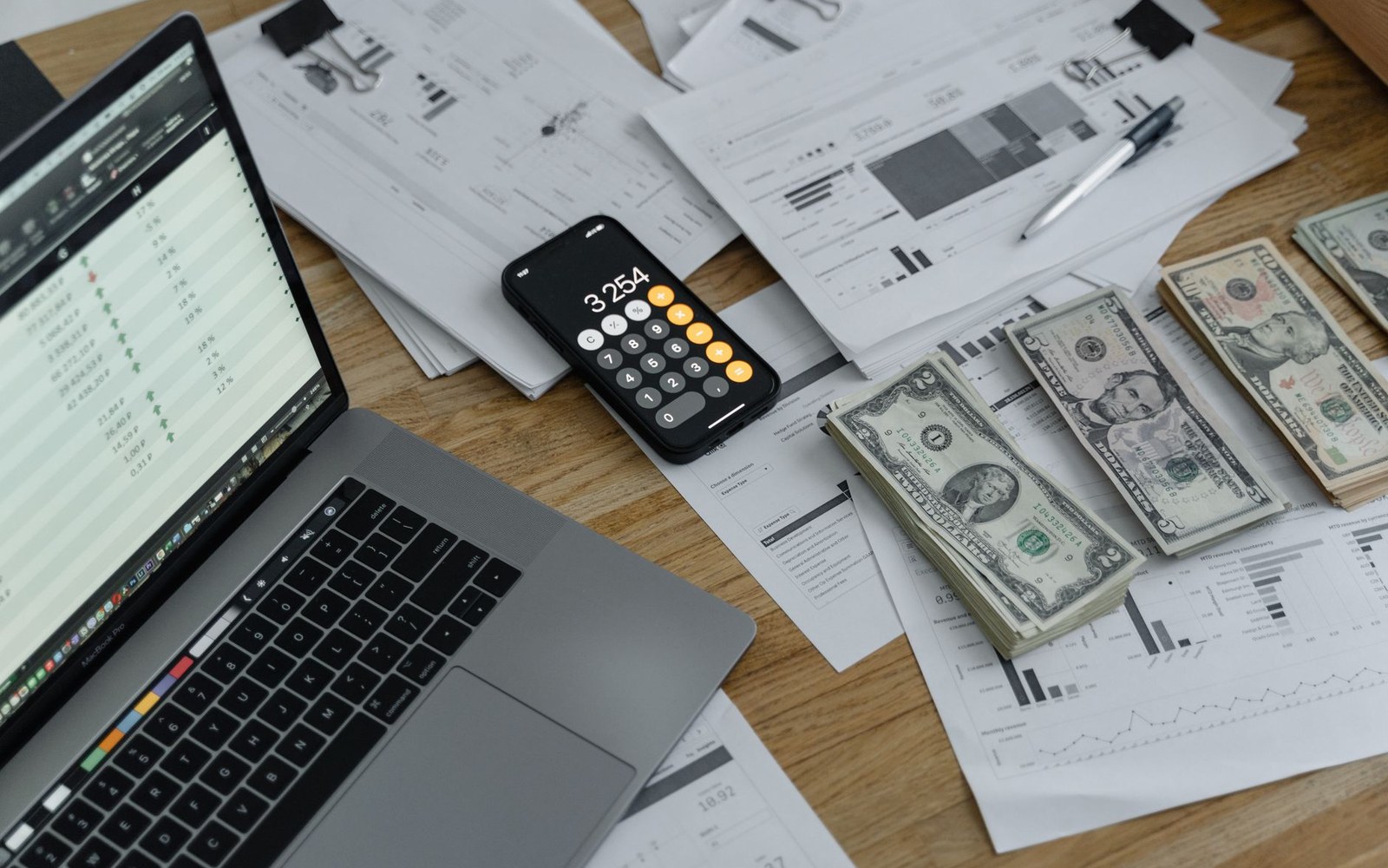 Laptop, phone calculator, cash and financial documents spread across a wooden desk