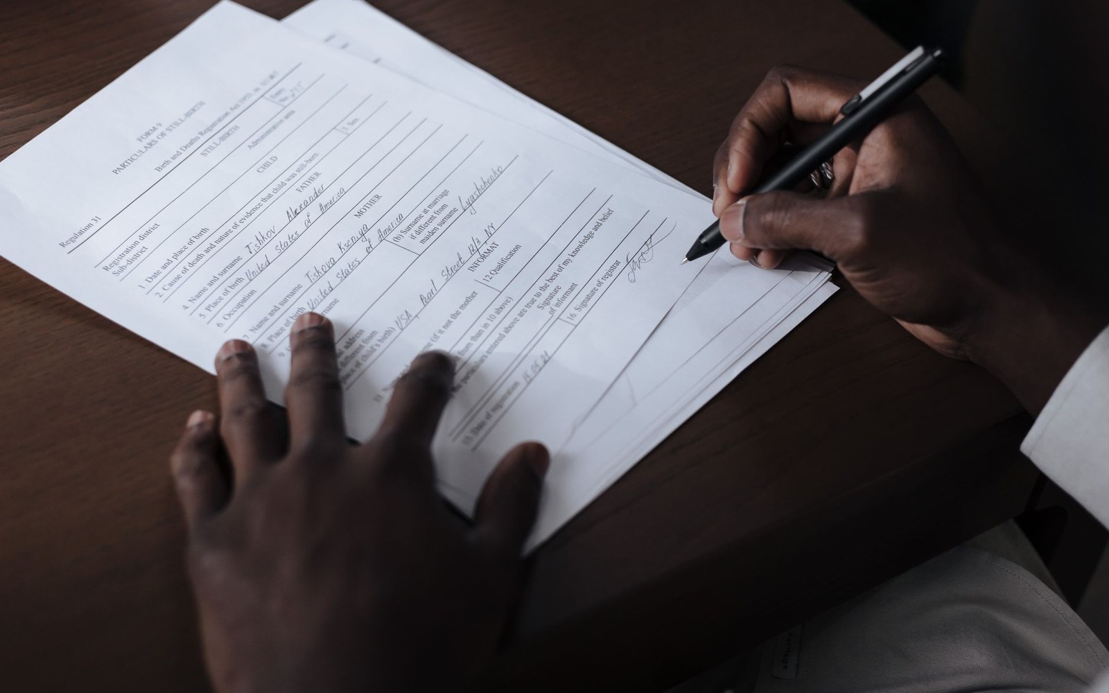 A person signs a printed form on a wooden desk with a pen